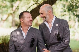 Two grooms sit on a bridge with dappled sun at the wedding venue after their ceremony at The Old BArn Esholt